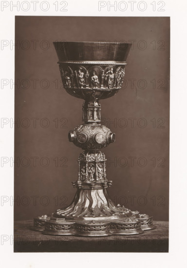 Chalice from a monastery in Tours, installed during the Exposition Rétrospective of 1873 in Tours, France, page, photomechanical print, maker: Gabriel Blaise, Tours, 1873, paper, Woodburytype, Woodburytypepie, height 190 mm x width 125 mm, temporary exhibition, 'Salon', chalice, Period, 1873-05-03 - 1873-05-25, A tall ornate metal chalice is centered against a plain dark background, showing a wide bowl with a smooth inner surface and a decorative band around its lower rim featuring small arched niches each containing a standing human figure in relief; below the bowl a rounded knop with circular medallions and foliate patterns connects to a columnar section decorated with miniature sculpted figures and architectural details, and the base flares into a patterned pedestal with flowing, wave-like ridges and an outer platform adorned with repeating ornamental motifs.