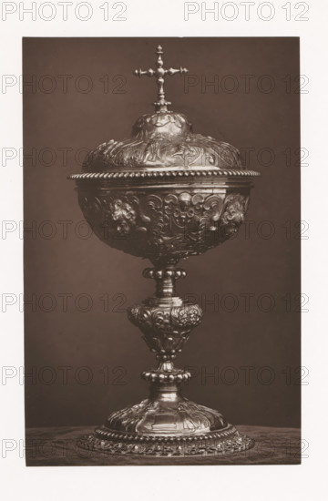 Ciborium of precious metal, installed during the Exposition Rétrospective of 1873 in Tours, France, page, photomechanical print, maker: Gabriel Blaise, Tours, 1873, paper, Woodburytype, Woodburytypepie, height 196 mm x width 126 mm, temporary exhibition, 'Salon', ciborium, Period, 1873-05-03 - 1873-05-25, A tall ornate chalice with a hinged domed lid topped by a small cross sits against a plain dark background, the body and stem covered in intricate raised floral and scrollwork decorations and small sculpted faces, the base broad and heavily ornamented with layered rosettes and beaded edges, the metal surface showing highlights and shadows that emphasize the embossed details.