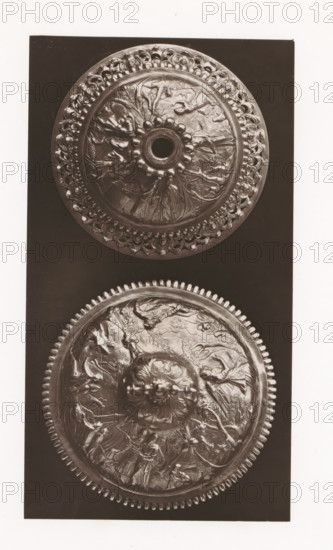 Two ciborium lids, installed during the 1873 Exposition Rétrospective in Tours, France, page, photomechanical print, maker: Gabriel Blaise, Tours, 1873, paper, Woodburytype, Woodburytypepie, height 202 mm x width 111 mm, temporary exhibition, 'Salon', ciborium, Period, 1873-05-03 - 1873-05-25, A vertical photograph shows two round, ornate metal lids or medallions set against a dark rectangular background and surrounded by a light border. Each round piece features a raised central knob with petal-like protrusions around a central opening on the upper one and a carved floral boss on the lower one. Both discs display intricate relief scenes with human figures, animals, drapery, and flowing organic motifs radiating outward, and each rim is embellished with repeating decorative elements; the upper rim has a scalloped, bead-like pattern while the lower rim shows a toothed, gear-like border. The metal surfaces exhibit highlights and shadows that emphasize the embossed details and textured wor...