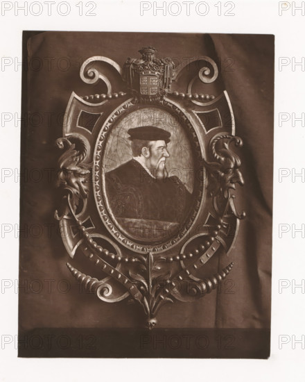 Portrait of Anne de Montmorency in a decorated frame, mounted during the Exposition Rétrospective of 1873 in Tours, France, page, photomechanical print, maker: Gabriel Blaise, Tours, 1873, paper, Woodburytype, Woodburytypepie, height 170 mm x width 131 mm, Person, Anne de Montmorency, historical persons, picture, painting, temporary exhibition, 'Salon', Period, 1873-05-03 - 1873-05-25, A framed oval portrait of a bearded man in profile wearing a cap and ornate clothing sits at the center of an elaborate carved wooden surround. The surround features scrolling volutes, beaded garlands, and symmetrical decorative elements including stylized faces with curled horns on either side. Above the oval is a crest with draped fabric and ornamental detailing, and below the frame are acanthus-like leaves and curved, ribbed scrolls ending in a small skull motif. The image has a sepia tone and the background shows a dark draped cloth behind the framed composition.
