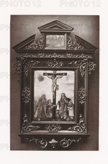 Enamel image of the crucifixion of Christ, displayed during the Exposition Rétrospective of 1873 in Tours, France, page, photomechanical print, maker: Gabriel Blaise, Tours, 1873, paper, Woodburytype, Woodburytypepie, height 193 mm x width 127 mm, picture, painting, temporary exhibition, 'Salon', crucified Christ, with particular persons under the cross, enamelling, applied arts, Period, 1873-05-03 - 1873-05-25, A tall ornate rectangular wooden frame with carved scrollwork surrounds a painted scene of a crucifixion; the frame has a pediment at the top with a small recessed panel and decorative curving motifs on either side, layered molding and symmetrical carved flourishes running along the borders. The central painted panel shows a figure on a cross with an expressive posture, a radiant circular shape to one side and a smaller crescent shape on the other, and several robed figures gathered at the base of the cross in varied standing and kneeling poses. The frame displays corner and si...