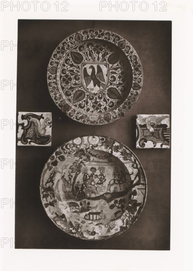 Two bowls and two earthenware tiles, displayed during the Exposition Rétrospective of 1873 in Tours, France, page, photomechanical print, maker: Gabriel Blaise, Tours, 1873, paper, Woodburytype, Woodburytypepie, height 194 mm x width 132 mm, temporary exhibition, 'Salon', dish, plate, saucer, Period, 1873-05-03 - 1873-05-25, A vertical photograph shows two large decorative plates and two square tiles arranged on a dark background, the upper plate featuring an ornate floral border and a central shield with a stylized bird, the lower plate displaying a busy scene with many small figures and landscapes around its rim, and the two square tiles positioned between the plates each containing different ornamental motifs including a curled decorative shape on one and a winged creature on the other.