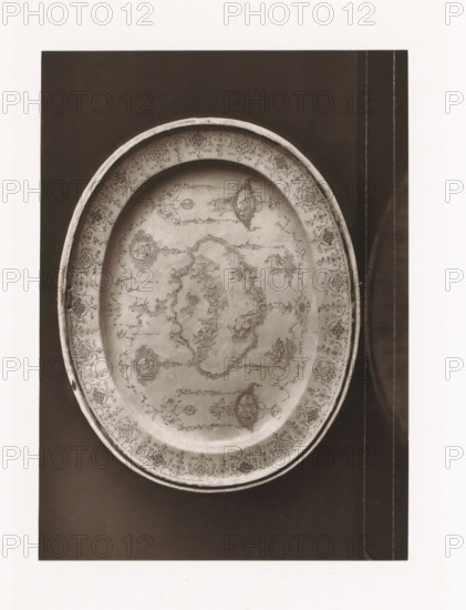 Small earthenware bowl from a monastery, displayed during the Exposition Rétrospective of 1873 in Tours, France, page, photomechanical print, maker: Gabriel Blaise, Tours, 1873, paper, Woodburytype, Woodburytypepie, height 165 mm x width 120 mm, temporary exhibition, 'Salon', dish, plate, saucer, Period, 1873-05-03 - 1873-05-25, An oval metal tray with an ornate etched design fills most of the image, showing a raised rim with a patterned border and an interior decorated with intricate floral and geometric motifs and symmetrical medallions; the tray rests on a dark rectangular surface with narrow lighter vertical lines near the right edge, and a white margin surrounds the dark background.