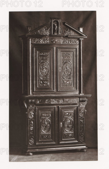 Cabinet from Château d'Anet, installed during the Exposition Rétrospective of 1873 in Tours, France, page, photomechanical print, maker: Gabriel Blaise, Tours, 1873, Gabriel Blaise, paper, Woodburytype, Woodburytypepie, height 197 mm x width 116 mm, temporary exhibition, 'Salon', ornamental cupboard (wood and other phytogenic material), Period, 1873-05-03 - 1873-05-25, A tall dark wooden cabinet with ornate carved panels and decorative pilasters, featuring upper and lower sections with framed doors, oval relief medallions depicting human figures, floral and scroll motifs, a triangular pediment at the top with a central niche holding a carved standing figure, carved horizontal friezes with reclining figures, fluted columns on either side, molded base and rounded feet, and a dark backdrop behind the cabinet.