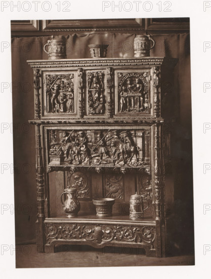 Sideboard in the style of Francis I of France, displayed during the Exposition Rétrospective of 1873 in Tours, France, page, photomechanical print, maker: Gabriel Blaise, Tours, 1873, paper, Woodburytype, Woodburytypie, height 179 mm x width 130 mm, temporary exhibition, 'Salon', ornamental cupboard (wood and other phytogenic material), Period, 1873-05-03 - 1873-05-25, A tall wooden cabinet with ornate carved panels and columns fills the frame, its upper section featuring three framed relief scenes set beneath a carved cornice, with a small pitcher and a lidded tankard placed on top and a small cup between them, the middle register showing a detailed horizontal relief of figures and horses within an architectural setting, and the lower shelving holding a decorated jug, a wide bowl, and a tall tankard, all set against a draped backdrop and photographed in monochrome.