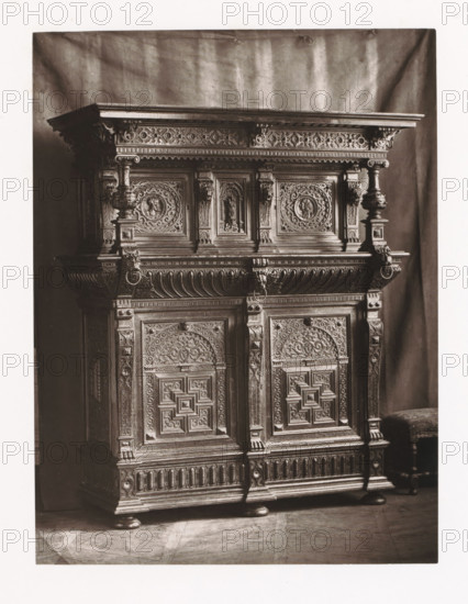 Flemish style sideboard, installed during the 1873 Exposition Rétrospective in Tours, France, page, photomechanical print, maker: Gabriel Blaise, Tours, 1873, paper, Woodburytype, Woodburytypepie, height 213 mm x width 161 mm, temporary exhibition, 'Salon', ornamental cupboard (wood and other phytogenic material), Period, 1873-05-03 - 1873-05-25, A large ornate wooden cabinet fills the frame, showing the upper section and doors with elaborate carved panels and decorative molding; the top has a flat projecting cornice with carved frieze beneath, supported by carved columns and brackets featuring rounded baluster shapes and carved lion head rings, while the door panels display intricate geometric and floral reliefs surrounded by carved borders and small columnar pilasters; the lower section shows carved vertical fluting and rounded feet resting on a patterned floor, and a small upholstered stool with turned legs is visible at the right edge against a draped backdrop.