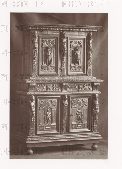 Cabinet in the style of Henry III of France, installed during the Exposition Rétrospective of 1873 in Tours, France, page, photomechanical print, maker: Gabriel Blaise, Tours, 1873, paper, Woodburytype, Woodburytypepie, height 184 mm x width 123 mm, temporary exhibition, 'Salon', ornamental cupboard (wood and other phytogenic material), Period, 1873-05-03 - 1873-05-25, The image shows a tall wooden cabinet with ornate carved panels and decorative molding, featuring sculpted human figures in recessed rectangular frames, fluted pilasters with carved faces and foliage, a projecting cornice with carved geometric and leaf patterns, a middle shelf with scrollwork and raised relief motifs, paneled doors with central standing figures, carved rosettes and acanthus leaves, and the cabinet resting on rounded bun feet on a wooden floor.