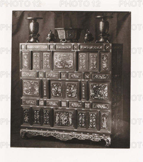 Cabinet from Annam, installed during the Exposition Rétrospective of 1873 in Tours, France, page, photomechanical print, maker: Gabriel Blaise, Tours, 1873, paper, Woodburytype, Woodburytypepie, height 178 mm x width 154 mm, temporary exhibition, 'Salon', ornamental cupboard (wood and other phytogenic material), Period, 1873-05-03 - 1873-05-25, A tall wooden cabinet fills the frame, its surface covered in numerous rectangular panels each decorated with intricate painted scenes and floral motifs; the cabinet rests on a carved base with geometric fretwork and curved feet. On top of the cabinet sit several metal vessels including two tall urns with wide flat lids positioned near the outer edges, a large square-shaped bowl with ornate relief work in the center, and two smaller lidded containers with sculpted figures on their lids flanking the central bowl. The background is a plain dark cloth that contrasts with the lighter tones of the cabinet and metal objects.