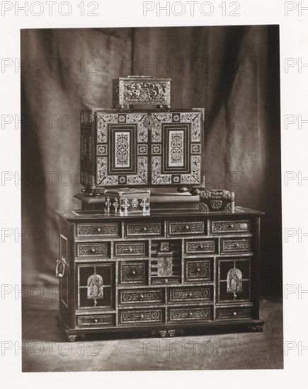 Two cabinets and three chests, displayed during the 1873 Exposition Rétrospective in Tours, France, page, photomechanical print, maker: Gabriel Blaise, Tours, 1873, paper, Woodburytype, Woodburytypepie, height 206 mm x width 156 mm, temporary exhibition, 'Salon', chest, coffer (wood and other phytogenic material), ornamental cupboard (wood and other phytogenic material), Period, 1873-05-03 - 1873-05-25, A photograph showing a multi-tiered ornate cabinet with intricate inlaid patterns and decorative panels placed against a draped fabric backdrop, the cabinet featuring a larger lower section with many small drawers and framed panels, a central open compartment with a carved figure, ornate metal ring handles on the sides, and raised feet, while a smaller decorated box sits atop the cabinet and an additional rectangular box with carved floral motifs rests above that, all finished with detailed filigree and contrasting light and dark surfaces.