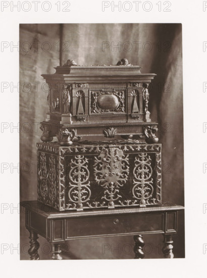 Two chests, displayed during the 1873 Exposition Rétrospective in Tours, France, page, photomechanical print, maker: Gabriel Blaise, Tours, 1873, paper, Woodburytype, Woodburytypepie, height 168 mm x width 117 mm, temporary exhibition, 'Salon', chest, coffer (wood and other phytogenic material), Period, 1873-05-03 - 1873-05-25, A photograph of a decorative chest displayed on a small wooden table, showing intricate carved patterns and ornamental details; the chest features ornate scrollwork panels with symmetrical floral and circular motifs on its sides, a patterned base with rounded feet, and a raised upper section adorned with sculpted figures, fluted columns, a central oval cartouche, and beaded molding; the table beneath has a drawer front, turned legs, and a polished finish, all set against a softly draped fabric backdrop.