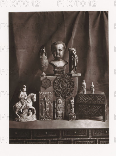 Various objects, displayed during the Exposition Rétrospective of 1873 in Tours, France, page, photomechanical print, maker: Gabriel Blaise, Tours, 1873, paper, Woodburytype, Woodburytypepie, height 165 mm x width 124 mm, temporary exhibition, 'Salon', Period, 1873-05-03 - 1873-05-25, A display of sculptural and carved objects arranged on a surface in front of a draped fabric backdrop includes a central bust of a child's head on a pedestal, flanked by an ornate vase with a narrow neck and a tall fragmented figure on a base, a round decorative medallion with elaborate foliage detailing mounted on a box, two small standing figurines on a raised box to the right, and a carved chest with geometric and floral patterns; below and in front are a mounted knight on horseback poised above a prone figure, several vertical carved panels depicting seated and standing figures and ornamental motifs, and a small column-like figure on a square base, all positioned on top of a richly decorated cabinet w...