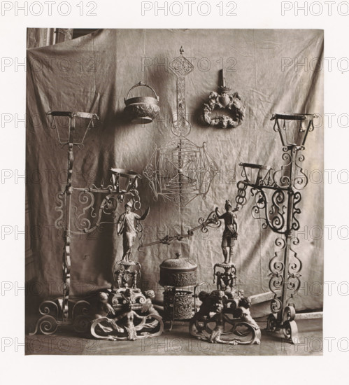 Various objects, displayed during the Exposition Rétrospective of 1873 in Tours, France, page, photomechanical print, maker: Gabriel Blaise, Tours, 1873, paper, Woodburytype, Woodburytypepie, height 181 mm x width 165 mm, temporary exhibition, 'Salon', Period, 1873-05-03 - 1873-05-25, A sepia-toned photograph shows a backdrop of draped fabric with decorative metal objects arranged in front of it, including ornate wrought iron stands with scrollwork on either side, a hanging bowl with embossed patterns suspended by a handle, a tall decorative metal panel with intricate openwork and a pointed finial, and a carved ornamental wreath with sculpted animal heads. In the lower area there are sculpted figurines mounted on bases, a lidded cylindrical container with embossed bands, multiple intricately carved supports featuring seated cherubic figures, and connecting metal rods and chains linking some of the elements together.