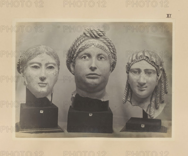 Three fragments of Egyptian sculptures, female heads, photograph, page, number: 'XI', c. 1890 - c. 1895, photographer, paper, gelatin silver print, developed gelatin silver print, height 157 mm x width 210 mm, sculpture, adult woman, head (human), Three sculpted stone heads are shown mounted on dark rectangular bases against a neutral background; the left head has short wavy hair, hollowed eyes, a narrow nose, and a small closed mouth with a smooth, worn surface; the central head features a wrapped turban with braided texture, prominent rounded cheeks, wide open eyes, and fuller lips with a smooth chin and neck fragment; the right head displays long braided hair draped over the shoulder, a rounded forehead, almond-shaped eyes, and a subtle smile with visible surface texture and small chips at the edges.