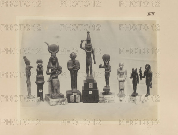 Nine Egyptian objects, including bronze sculptures of Mut, Anupt, Isis with Horis and Apis, photograph, page, number: 'XIII', c. 1890 - c. 1895, photographer, paper, gelatin silver print, developing gelatin silver print, height 158 mm x width 213 mm, sculpture, representations, Egyptian gods, demi-gods, heroes, etc. (with NAME), Horus, son of Osiris and Isis, Isis, the mother-goddess, Egyptian goddesses, demi-goddesses, heroines, A row of small dark statues and figurines stands on a flat surface against a light background, each mounted on individual pedestals of varying shapes and heights; the figures include humanlike forms with headdresses and seated postures, a figure holding a smaller figure, a standing figure with an arm raised and another with a circular element above its head, a taller central figure with an arm raised holding a horizontal object and a carved face, and several smaller standing figures with distinct headgear and limbs posed differently, all appearing in a uniform...