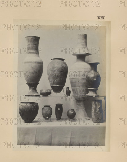 Eleven Egyptian vases, photograph, page, number: 'XIX', c. 1890 - c. 1895, photographer, paper, gelatin silver print, developing gelatin silver print, height 208 mm x width 155 mm, containers, A centered photograph mounted on a pale sheet shows a display of ceramic vessels arranged on shelves draped with fabric; the top shelf holds a variety of tall urns and amphorae with rounded bodies, flared rims, narrow necks, and pedestal bases in muted tones and worn surfaces, including a large bulbous vessel, a tall slender vase with a wide flaring neck, and a conical-necked jar, while the lower shelf shows smaller pots and jars of different shapes including globular and cylindrical forms, one with spiral decorative motifs and another with vertical leaf-like patterns, several supported on small metal stands; the overall image has a soft, slightly faded monochrome appearance and visible texture and wear on the ceramics and paper.