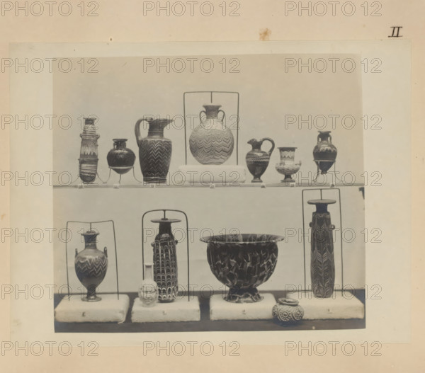 Eleven Egyptian vases and a cup and a pot, photograph, page, number: 'II', c. 1890 - c. 1895, photographer, paper, gelatin silver print, developed gelatin silver print, height 154 mm x width 205 mm, sculpture, container of glass: bottle, jar, vase, container of stone: stone vase, A framed photograph shows a display of various pottery pieces arranged on padded stands and metal supports against a plain backdrop; the visible vessels include tall slender amphorae with patterned surfaces, handled jugs with spouts and zigzag or wave-like designs, a large bowl with a mottled or marbled interior, small rounded jars with ornate surface decoration, and a sculpted head object placed on one of the stands, all in muted tones and photographed in a museum-style arrangement.