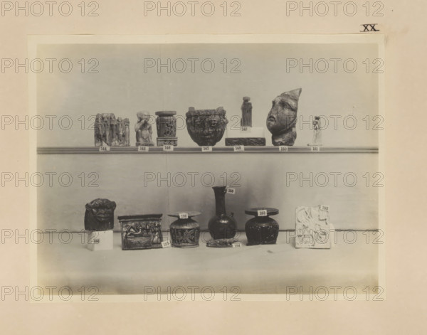 Fourteen Egyptian objects, including a sculpture of Harpocrates and four cabbage pots, photograph, page, number: 'XX', c. 1890 - c. 1895, photographer, paper, gelatin silver print, developing gelatin silver print, height 160 mm x width 212 mm, sculpture, containers, representations, Egyptian gods, demi-gods, heroes, etc. (HARPOCRATES), A shelf display of various small sculptural objects and vessels arranged in two rows against a plain background, including small carved figures, a fragmented human-like head, cylindrical and rounded pots with decorative relief, a carved rectangular box, a lion-like carved block, a tall narrow-necked vase, and a rectangular carved slab, all shown in monochrome with subtle shadows on the shelf and base.