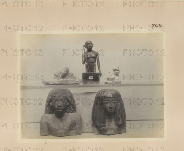 Five Egyptian objects, including two busts, photograph, page, number: 'XXIII', c. 1890 - c. 1895, photographer, paper, gelatin silver print, developing gelatin silver print, height 159 mm x width 214 mm, sculpture, adult woman, adult man, A faded photograph mounted on a beige background shows a display of carved stone figures on a shelf, with a smaller standing figure holding a staff placed above two seated busts below; the seated busts have textured hair or wigs and detailed facial features, while small fragmentary sculptures flank the standing figure on the upper shelf.