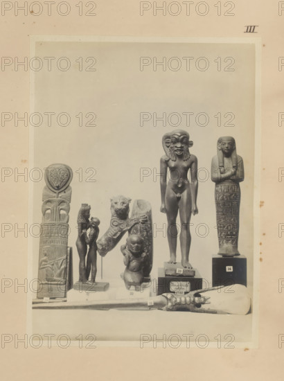 Seven Egyptian objects, including a bronze sculpture of Isis with Horus, a bronze sculpture of Harpocrates with the head of Bes and a wooden ushabti, photograph, page, number: 'III', c. 1890 - c. 1895, photographer, paper, gelatin silver print, developed gelatin silver print, height 212 mm x width 157 mm, sculpture, Horus, son of Osiris and Isis, Isis, the mother-goddess, representations, Egyptian gods, demi-gods, heroes, etc. (BES), A sepia-toned photograph shows a display of small statues and carved objects arranged on pedestals and a flat surface. From left to right there is a tall vertical carved pillar topped with a round emblem, a slender figurine of a standing pair with one figure slightly smaller and facing the taller figure, a sculpted animal head mounted on a fragment of carved stone beside a small rounded figure with a childlike form, a taller nude male figure with an exaggerated facial mask standing on a square base, and a mummiform figure with arms crossed and detailed pat...