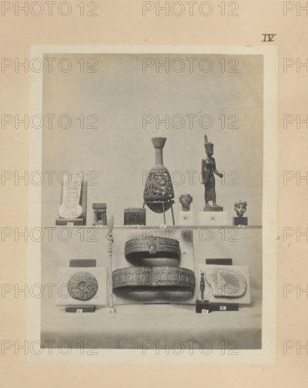 Twelve Egyptian objects, including a bronze sculpture of Neith, two gold bracelets and a gold sculpture of Hathor, photograph, page, number: 'IV', c. 1890 - c. 1895, photographer, paper, gelatin silver print, developing gelatin silver print, height 203 mm x width 158 mm, sculpture, bracelet, wristlet, representations, Egyptian goddesses, demi-goddesses, heroines, etc. (NEITH), A framed black-and-white photograph of a display shelf filled with small artifacts, centered on a tall decorative vessel with a narrow neck and flared top, flanked by a standing figurine wearing a tall headdress and a sculpted head on simple rectangular plinths, with a carved rectangular tablet and several small box-like objects on the left side, and a small sculpted head on a plinth on the right; below the shelf are two ornate circular boxes stacked above a rounded base, a tall slender staff with a figure near its top, a round carved disk mounted on a cushion, a small standing figurine on a square base, and a re...