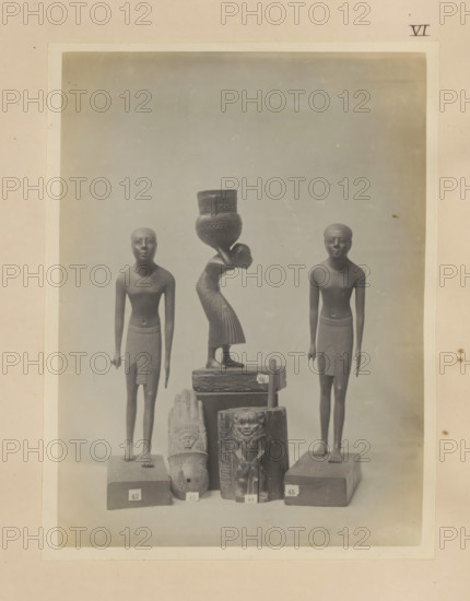 Five Egyptian objects, including two sculptures of boys and a wooden kohl pot with the image of Bes, photograph, page, number: 'VI', c. 1890 - c. 1895, photographer, paper, gelatin silver print, developed gelatin silver print, height 212 mm x width 161 mm, sculpture, youth, adolescent, wooden container: barrel, cask, bucket, representations, Egyptian gods, demi-gods, heroes, etc. (BES), A framed photograph shows a group of carved figures arranged on a flat surface against a neutral backdrop, including two standing male figures with short hair and wrapped skirts positioned on either side, a central female figure bent forward carrying a large decorated vessel on her head, several smaller carved objects and blocks with relief faces and patterns stacked in front, and a pale border surrounding the image with a small mark near the top right.