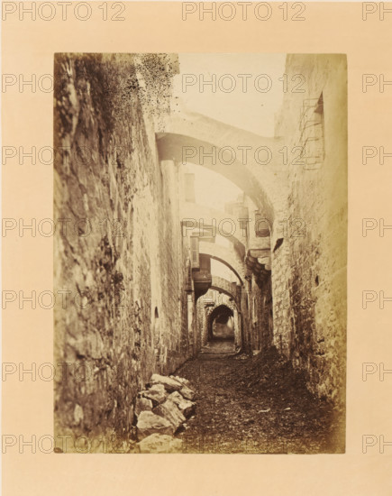 View into the Via Dolorosa, photograph, page, photographer: Horatio Herbert Kitchener, Via Dolorosa, c. 1870 - c. 1875, Jerusalem, paper, albumen print, height 201 mm x width 147 mm, street, arch, archivolt, architecture, A narrow stone passageway framed by rough textured walls on both sides extends into the distance, with a series of curved stone arches spanning overhead and casting soft shadows. The ground is uneven and covered with small stones and dirt, and a pile of larger rubble rests along the left base of the wall. Small, shuttered window openings and protruding stone corbels line the walls, and a metal window grate is visible on the right wall. The photograph has a faded, sepia tone and shows wear and spotting near the top.