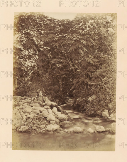 View of Elisha's Fountain in Jericho, photograph, page, photographer: Horatio Herbert Kitchener, Jericho, c. 1870 - c. 1875, Horatio Herbert Kitchener, photographer, Palestine, West Bank, paper, albumen print, height 200 mm x width 151 mm, spring, source of river or stream, A sepia-toned photograph showing a rocky stream flowing from the middle toward the bottom foreground, with smooth water over and between rounded stones; dense foliage and tall leafy plants rise behind the stream, creating a dark textured backdrop of leaves and branches; a person wearing a hat and coat reclines on a pile of rocks at the stream edge, leaning on one elbow and looking slightly upward, with legs extended and one knee bent; shrubs and grasses grow along the stream banks and some branches and sticks lie among the stones.