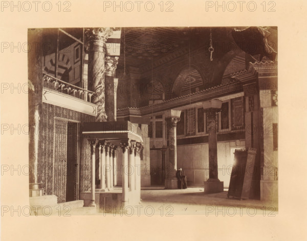 Interior of the Dome of the Rock on the Temple Mount, photograph, page, photographer: Horatio Herbert Kitchener, Dome of the Rock, c. 1870 - c. 1875, Jerusalem, paper, albumen print, height 155 mm x width 206 mm, temple, shrine, Islam, Mohammedanism, A sepia photograph of an interior space showing tall decorative columns with ornate capitals and marble-like surfaces, arched ceilings with patterned coffers, and a row of slender columns supporting a balcony or canopy with carved details; patterned wall coverings and carved panels line one side, a large rectangular column or pulpit structure stands in the foreground, and a person seated on a bench wearing dark clothing and a head covering is visible near the back beneath the arches.