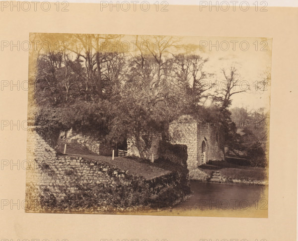 Fortress gate of Leeds Castle, photograph, page, inscription: 'No.3.', photographer: John Cruttenden, Leeds Castle, c. 1864 - in or before 1869, Leeds, paper, albumen print, height 153 mm x width 204 mm, landscape with tower or castle, castle, gate, entrance, A sepia-toned photograph shows a stone ruin with an arched doorway partially obscured by foliage, situated beside a body of water. Dense trees with bare and leafy branches rise behind and above the ruin, their trunks and tangled limbs forming a dark canopy. A stone wall and terraced grassy areas lead down toward the water, with short fence posts and a wooden gate visible on the grassy path. The surface of the water appears calm and reflective near the stone edge. The image is bordered by a plain light-colored mat.