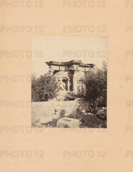View of the Temple of Baal at Baalbek, photograph, page, annotation: 'Temple of Baal at Baalbek', photographer: Francis Frith, Baalbek, photographer: Frank Mason Good, c. 1870 - in or before 1875, paper, albumen print, height 101 mm x width 82 mm, Photo after page 40., temple, shrine, Roman religion, A faded rectangular photograph centered on a wider beige background shows stone ruins partially obscured by dense foliage; the ruins include a carved stone facade with ornate horizontal cornices and arched openings, weathered blocks and scattered stones in the foreground, a low rough stone wall, and leafy trees growing around and behind the structure, with shadows and textured vegetation filling the lower portion of the image.