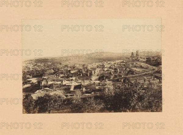 View of Al-Eizariya or Bethany, photograph, page, annotation: 'Bethany', photographer: Francis Frith, Al-Eizariya, photographer: Frank Mason Good, c. 1870 - in or before 1875, Palestine, paper, albumen print, height 104 mm x width 154 mm, city-view in general, 'veduta', landscape with ruins, A wide, faded monochrome photograph mounted on a pale background shows a hillside landscape with a cluster of stone ruins and low buildings spreading across terraces. The foreground contains darker, leafy vegetation and scattered trees, transitioning to rough stone walls, fragmented structures, and archways among uneven ground. Further back, more ruined walls and partial towers rise near a low ridge, and open fields and rolling hills extend toward the horizon under a blank, light sky.