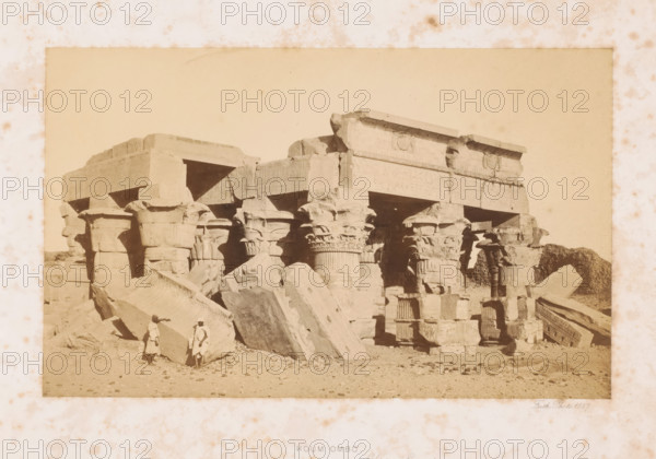 Two unknown men at the ruins of the temple of Kom Ombo, Koum Ombo, page, This photo is part of a cover with 7 pages of albumen prints., inscription: 'Upper Egypt', photographer: Francis Frith, Kom Ombo, 1857, North Africa, paper, albumen print, height 142 mm x width 231 mm, Person, historical persons, ruin of a building, architecture, temple, shrine, Egyptian religion, A sepia-toned photograph shows a ruined stone structure with large, weathered blocks and carved columns; the columns have ornate capitals and fluted shafts, and some blocks lie toppled in front of the structure. Two men in light clothing and head coverings stand among the fallen stones near the foreground. The sky above the ruins appears pale and empty, and the ground in front of the structure is sandy with scattered small rocks. The image is framed by a lighter border with spotting and discoloration.