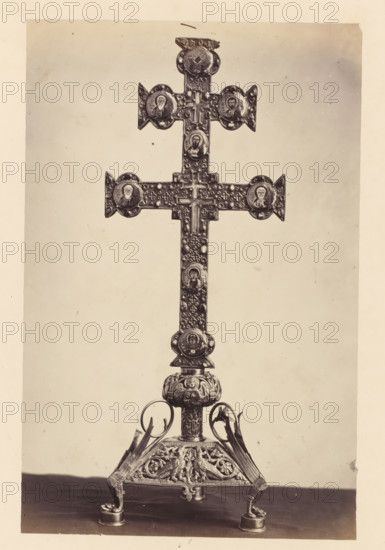 Golden cross with relic from the collection of the Soeurs de Notre-Dame de Namur, displayed at an exhibition of religious objects from the Middle Ages and Renaissance in 1864 in Mechelen, photograph, page, number: '1', photographer: Joseph Maes, Mechelen, 1864, paper, albumen print, height 269 mm x width 175 mm, relics (and reliquaries), piece of sculpture (precious metals, arts), Period, A tall ornate metal cross mounted on an elaborately decorated stand fills the frame, with a vertical shaft and a transverse beam forming a cross shape adorned with circular medallions featuring portrait-like images and textured decorative elements; the vertical shaft shows repeated small raised motifs and additional medallions, and the top and ends of the horizontal arm have pointed triangular extensions. The stand beneath the cross is highly detailed with sculpted figures and winged motifs, curved supports rising to meet the central column, and small rounded feet resting on a flat surface. The backgr...