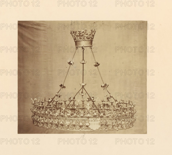 Two metal candlesticks with bases, displayed at an exhibition of religious objects from the Middle Ages and Renaissance in 1864 in Mechelen, photograph, page, number: '17 - 18', photographer: Joseph Maes, Mechelen, 1864, paper, albumen print, height 258 mm x width 196 mm, chandelier, candelabrum, Period, A large ornate metal crown suspended by slender rods is visible against a plain backdrop, the crown featuring a circular base with repeated floral and arched decorative motifs, numerous small cup-like holders around the rim, vertical supports rising to a narrower crown-like top with intricate openwork and fleur-de-lis shaped elements, and decorative rings and curved connectors along the supports; the image has a wide margin of blank paper surrounding the photographed crown.