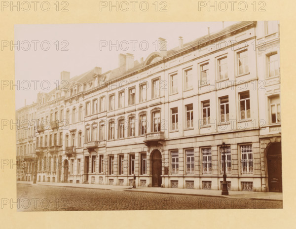 Street view in Brussels, photograph, page, c. 1890 - in or before 1895, photographer, paper, daylight collodion silver print, height 158 mm x width 224 mm, urban housing, street (city(-scape) with figures, staffage), Brussels, A sepia-toned photograph of a long row of multi-story urban buildings with ornate facades, tall rectangular windows, decorative molding and small balconies, a cobblestone street in front, a sidewalk with a single standing person near a building entrance, a streetlamp on the sidewalk, wrought-iron window grilles on the ground floor, chimneys and roof details visible along the roofline, and a light border surrounding the photograph.