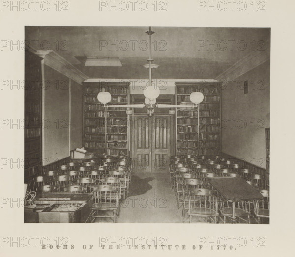 Rooms of the Institute of 1770, Interior of the Institute of 1770 of Harvard University, page, photomechanical print, c. 1870 - in or before 1875, paper, collotype, photoprint, height 164 mm x width 205 mm, university building, college, club (students), North America, Massachusetts, Cambridge (MA), Harvard College, A sepia-toned interior photograph showing a room with rows of wooden chairs arranged facing a central double door flanked by tall bookshelves filled with books, overhead light fixtures with round globe shades suspended from the ceiling, a long table visible at the right side with chairs around it, a smaller table with stacked books on the left, crown molding around the ceiling, and a patterned carpeted floor.