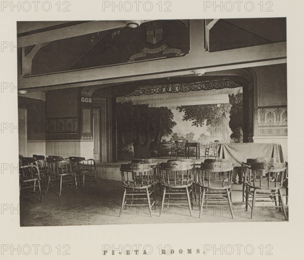 Theatre of the Harvard Pi Eta Society, Pi-Eta rooms, page, photomechanical print, c. 1870 - in or before 1875, paper, collotype, photoprint, height 160 mm x width 210 mm, university building, college, club (students), North America, Massachusetts, Cambridge (MA), Harvard College, A sepia-toned interior photograph showing a small stage with a painted backdrop of trees and a landscape, framed by decorative arches and trim, with two wooden chairs and a small table on the stage and a draped cloth covering a portion of the stage to the right; in front of the stage rows of wooden spindle-back chairs face forward, some grouped around small round tables, and wooden flooring is visible beneath; the ceiling above the stage has exposed beams and a mounted light fixture, and the walls feature patterned friezes and paneling.