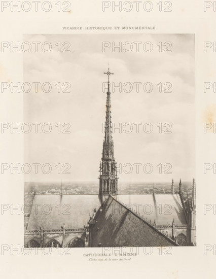 Tower on Notre-Dame of Amiens, page, photomechanical print, inscription: 'Cathédrale d'Amiens, Flèche oue de la tour de Nord', cliché maker: Paul Dujardin, c. 1888 - in or before 1893, Paul Dujardin, photographer, cliché maker, paper, collotype, photoprint, height 210 mm x width 170 mm, parts of church exterior and annexes: tower, Amiens, Notre-Dame d'Amiens, A sepia-toned photograph showing the top portion of a Gothic cathedral with a tall ornate spire rising from the roof, the spire topped by a decorative cross and featuring intricate tracery and sculptural details; the steep sloping roof planes of the cathedral flank the spire, with visible ridge lines, gutters, and rows of pinnacles along the roof edges; the image includes a distant hazy cityscape on the horizon below the roofline and a cloudy sky filling the upper portion of the frame; the overall image appears aged with subtle stains and toning around the paper margins.
