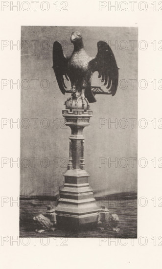 Sculpture of an eagle on a pedestal from the Basilica of Saint Martin in Halle, displayed at an exhibition of religious objects from the Middle Ages and Renaissance in 1864 in Mechelen, page, photomechanical print, number: '-49', number: 'I ...', number: '1', photographer: Joseph Maes, Mechelen, clichémaker: Simonau & Toovey, Antwerp, 1864 - in or before 1866, paper, photolithography, height 238 mm x width 132 mm, predatory birds: eagle, piece of sculpture, reproduction of a piece of sculpture (metals, arts), Period, 1864, A tall sculptural stand topped by a large eagle with outstretched wings perched on a rounded base, the eagle showing detailed feather texture and a forward-facing head, the stand beneath it featuring a multi-tiered column with ornamental moldings and carved panels, and four small lion figures positioned near the base on a flat surface; the scene appears in monochrome with a plain backdrop and visible grain from the photographic print.