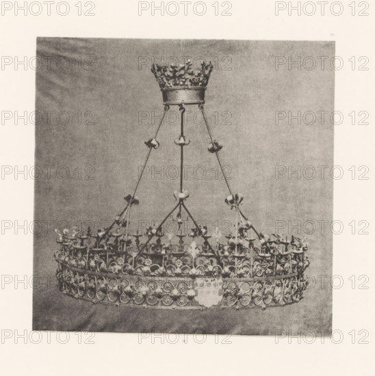 Wrought-iron chandelier from St. Peter's Church in Bastogne, displayed at an exhibition on religious objects from the Middle Ages and Renaissance in 1864 in Mechelen, page, photomechanical print, number: '-56', number: '16', photographer: Joseph Maes, Mechelen, clichémaker: Simonau & Toovey, Antwerp, 1864 - in or before 1866, Joseph Maes, Simonau & Toovey, photographer, clichémaker, Mechelen (heerlijkheid), paper, photolithography, height 168 mm x width 169 mm, chandelier, candelabrum, Period, 1864, A grayscale photograph of an ornate metal crown suspended by thin chains from a decorative circular support, showing a detailed upper band with repeating circular motifs and floral elements, small bowls or candle holders attached to the vertical supports, a smaller crown-like piece at the top with a textured leafy rim, and a plain backdrop behind the suspended object.