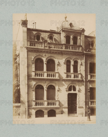Façade of a building in Paris, photograph, This photo is part of a cover with 35 photos and a title page., name: 'xxx Architecte', number: '12', photographer: Lampué et Cie, Paris, c. 1875 - in or before c. 1880, paper, albumen print, height 221 mm x width 165 mm, urban housing, façade (of house or building), A sepia-toned photograph of a multi-story stone building with arched windows and decorative stone balustrades; the facade shows rows of paired arched windows with shutters and ornamental columns between them, a central entrance recessed beneath an arched surround, and smaller arched basement openings with iron grilles; the uppermost level features rounded dormer windows set into a tiled mansard roof and a small domed roof section with a circular opening, while the roofline includes chimneys and small pipes; a cast-iron street lamp stands on the sidewalk near the building, and the image is bordered by a textured pale background.