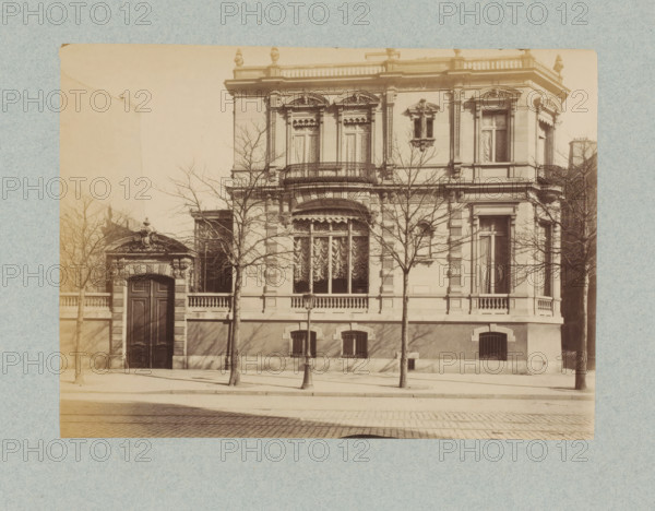Façade of a villa in Paris, designed by Jules Février, photograph, This photo is part of a cover with 35 photos and a title page., name: 'Février Architecte', number: '14', photographer: Lampué et Cie, Paris, c. 1875 - in or before c. 1880, paper, albumen print, height 165 mm x width 225 mm, detached house in city, façade (of house or building), A sepia-toned photograph of a large ornate building façade with decorative stonework and tall windows partially covered by patterned curtains, a curved balcony with wrought-iron railing above a bay window with swagged drapery, fluted pilasters and ornamental pediments above upper windows, a grand arched wooden doorway set in a small ornate stone gateway at one side, leafless trees planted along the sidewalk whose branches cast shadows on the façade, a lamppost on the pavement in front, small barred basement windows at street level, and a cobblestone street in the foreground.