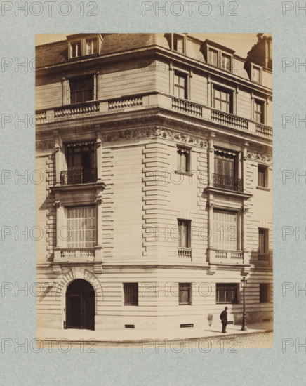 Corner of a building in Paris, photograph, This photo is part of a cover with 35 photos and a title page., name: 'Escalier Architecte', number: '18', photographer: Lampué et Cie, Paris, c. 1875 - in or before c. 1880, paper, albumen print, height 221 mm x width 165 mm, urban housing, A sepia-toned photograph of a multi-story corner building with ornate stonework and decorative cornices, featuring tall windows with shutters and iron balconies, a rounded arched entrance at street level, rusticated horizontal banding on the façade, carved frieze details below the upper balcony, dormer windows in the roof, a streetlamp near the corner, and a man standing on the sidewalk beside the lamp casting a shadow on the wall.