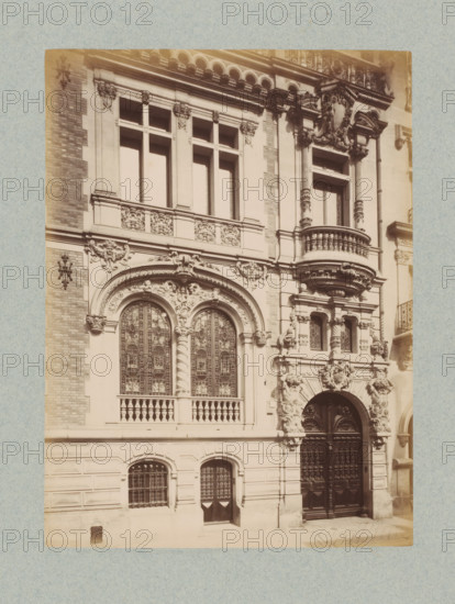 Façade of a residential house in Paris, photograph, This photo is part of a cover with 35 photos and a title page., name: 'E...ld .elterie', number: '22', photographer: Lampué et Cie, Paris, c. 1875 - in or before c. 1880, paper, albumen print, height 225 mm x width 166 mm, urban housing, façade (of house or building), A sepia-toned photograph of an ornate building façade showing carved stonework, decorative columns, balustrades, arched windows with patterned glass, and an elaborate double doorway with ornate panels and sculpted surround; there are sculpted faces and floral motifs above and around the arches, a rounded projecting balcony with turned balusters, rectangular windows set in decorative frames, textured brickwork at the left edge, and a stone sidewalk in front.