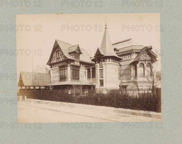 Villa in Paris, designed by Stephen Sauvestre, photograph, This photo is part of a cover with 35 photos and a title page., number: '34', name: 'Sauvestre Architecte', photographer: Lampué et Cie, Paris, c. 1875 - in or before c. 1880, paper, albumen print, height 166 mm x width 219 mm, detached house in city, A sepia-toned photograph of a large ornate building with steep gabled roofs, decorative half-timbered façades, a conical turret with a finial, arched windows, and masonry walls; a wrought-iron fence and a hedge run along the front, leafless trees stand near the building, and a paved street is visible in the foreground.