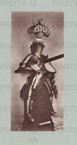 15th-century armor for a cavalryman from the army of Charles VII of France, from the collection of the Musée d'Artillerie in Paris, photomechanical print, This print is part of a cover with 36 prints, a title page and a list of images., check stamp: 'PHOTOGLYPTIE, LEMERCIER ET Cie, PARIS', Paris, cliché maker: Lemercier & Cie., c. 1877 - in or before 1882, Lemercier & Cie., photographer, cliché maker, paper, Woodburytype, Woodburytypie, height 266 mm x width 118 mm, armour, A full suit of polished plate armor displayed on a wooden stand, shown from a side angle with a lance held horizontally through a rounded shoulder plate, a rounded closed helmet with a decorative wide-brimmed crest topped by a small sculpted animal and horn-like projections, a cloth-lined collar visible beneath the helmet, articulated pauldrons, a segmented fauld and tassets over the hips, articulated cuisses and greaves on the legs, metal sabatons on the feet, and ornamental fluting and rivets on several pieces; th...