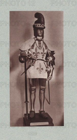 15th-century armor with various thrusting weapons from the army of Louis XI of France, from the collection of the Musée d'Artillerie in Paris, photomechanical print, This print is part of a cover with 36 prints, a title page and a list of images., check stamp: 'PHOTOGLYPTIE, LEMERCIER ET Cie, PARIS', Paris, cliché maker: Lemercier & Cie., c. 1877 - in or before 1882, Lemercier & Cie., photographer, cliché maker, paper, Woodburytype, Woodburytypie, height 278 mm x width 129 mm, hacking and thrusting weapons: sword, armour, A sepia-toned full-length photograph of a mannequin or statue dressed in medieval-style armor and a decorative surcoat, standing on a wooden plinth; the figure wears a helmet with a tall feather plume and a jeweled band, a layered shoulder cape over the surcoat, and a chain around the neck, with stylized fleur-de-lis motifs visible on the front of the surcoat; the arms show articulated plate armor and gauntlets, one hand resting on a long pole weapon and the other hol...