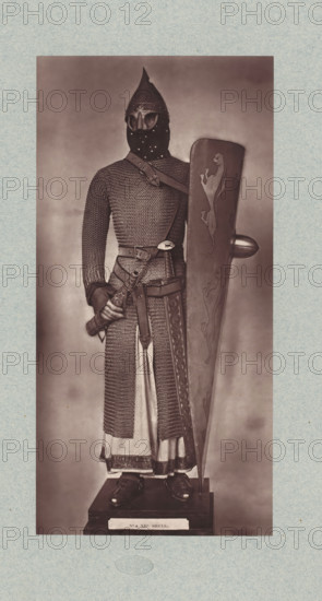 12th-century armor from the army of Louis VI of France, from the collection of the Musée d'Artillerie in Paris, photomechanical print, This print is part of a cover with 36 prints, a title page and a list of images., check stamp: 'PHOTOGLYPTIE, LEMERCIER ET Cie, PARIS', Paris, cliché maker: Lemercier & Cie., c. 1877 - in or before 1882, Lemercier & Cie., photographer, cliché maker, paper, Woodburytype, Woodburytypie, height 275 mm x width 135 mm, armour, helved weapons, polearms (for striking, hacking, thrusting): lance, A full-length sepia-toned photograph shows a person standing on a pedestal wearing medieval-style armor including a conical helmet with a pointed top and a face-covering mask with eye openings, a long chainmail hauberk extending down over a tunic, and gauntleted hands; a broad belt and cross-body strap with decorative buckles secure a sheathed sword at the waist, and one hand grasps the sword hilt while the other rests behind a tall kite-shaped shield decorated with st...
