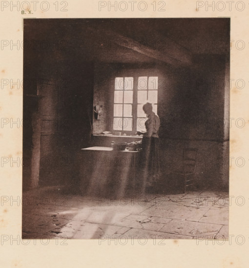 Woman at the window, page, photomechanical print, inscription: 'Interior by G. Marissiaux', inscription: 'BX', photographer: Gustave Marissiaux, cliché maker: anonymous, c. 1900 - in or before 1905, paper, two-color autotype, height 132 mm x width 123 mm, adult woman, interior of the house, A dark interior scene shows a woman standing by a multi-pane window with light streaming through, a table beneath the window holding small objects, a high ceiling with visible beams, a tiled floor with cracks and worn areas, and strong contrasts between illuminated and shadowed areas.