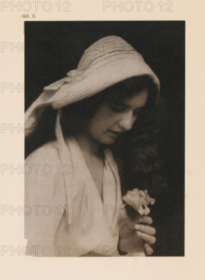 Portrait of an unknown woman with an iris, page, photomechanical print, inscription: 'GB. X.', inscription: 'The Iris by CF Juston', photographer: CF Juston, cliché maker: anonymous, c. 1900 - in or before 1905, paper, two-color autotype, height 149 mm x width 101 mm, historical persons - BB - woman, flowers: iris, A sepia-toned portrait of a young woman shown in profile against a dark background, wearing a textured headscarf that drapes over her hair and shoulders, her eyes cast downward and her expression neutral, her long curly hair visible beneath the scarf, a light-colored loose garment covering her upper body, and her hands gently holding a small cluster of light flowers near her chest.